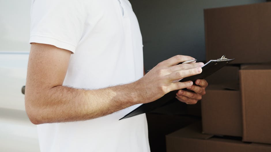 Close-up of a person wearing a white T-shirt, holding a black clipboard with both hands, in a home environment with cardboard boxes stacked nearby. The individual appears to be inspecting or planning a home relocation, with the background showing a dark interior wall and part of a shelving unit or additional boxes. The scene suggests involvement in packing, moving logistics, or inventory checking as part of a household furniture transport or removal process managed by Man With a Van Kingsbury, in preparation for a house move, with additional packing materials such as plastic wrap and cardboard visible. The lighting is neutral, highlighting the focus on the person's hands and the clipboard, with the context emphasizing professional removals and moving services.