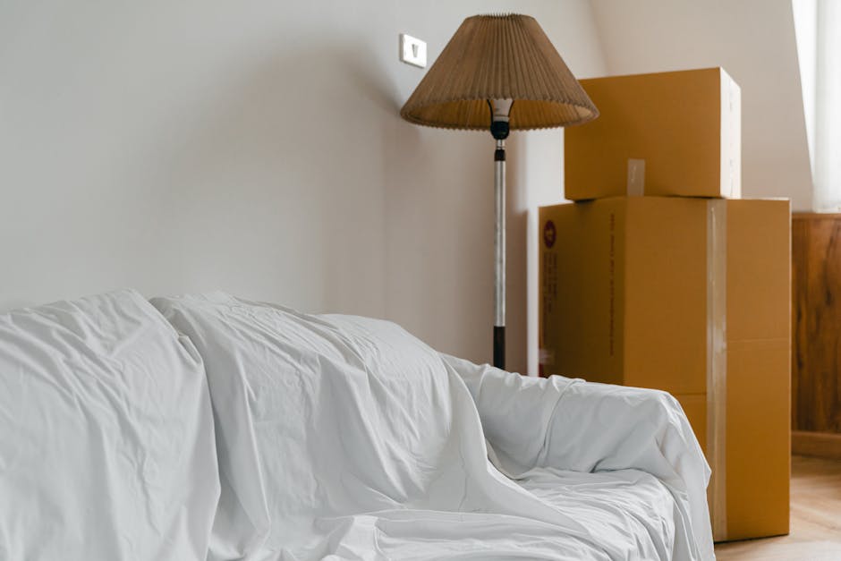 Inside a room during a home relocation process, a white sofa covered with a protective sheet is positioned near the left side of the image. Behind the sofa, three cardboard boxes of varying sizes are stacked, secured with clear packing tape. A tall, standing floor lamp with a beige pleated lampshade is situated to the right of the boxes. The boxes rest on a wooden sideboard or cabinet located against the wall, which is painted in a neutral, light colour. The area appears well-lit, with natural light coming from a window partly visible on the right side of the image. The scene reflects a typical moving environment, possibly during packing or loading for transport by a professional removals service such as Man With a Van Kingsbury, highlighting furniture, packing materials, and the logistical process involved in a house move within Kingsbury.