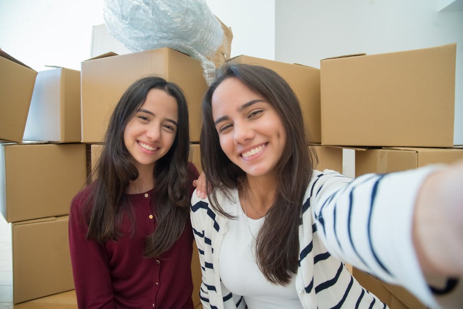 Two smiling women with dark brown hair taking a selfie inside a property during a house removal process. They are standing in front of a stack of cardboard boxes and packing materials, including a pla