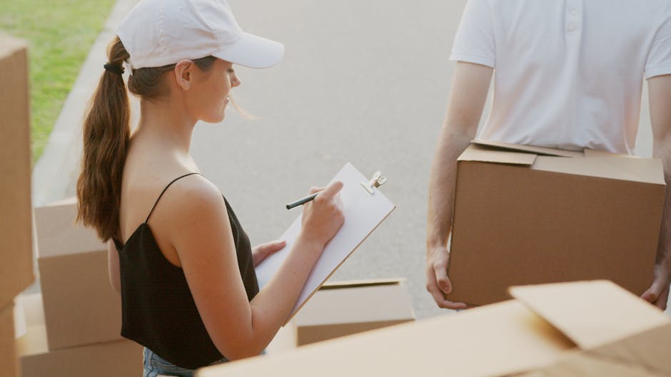 Two smiling women with dark brown hair taking a selfie inside a property during a house removal process. They are standing in front of a stack of cardboard boxes and packing materials, including a pla