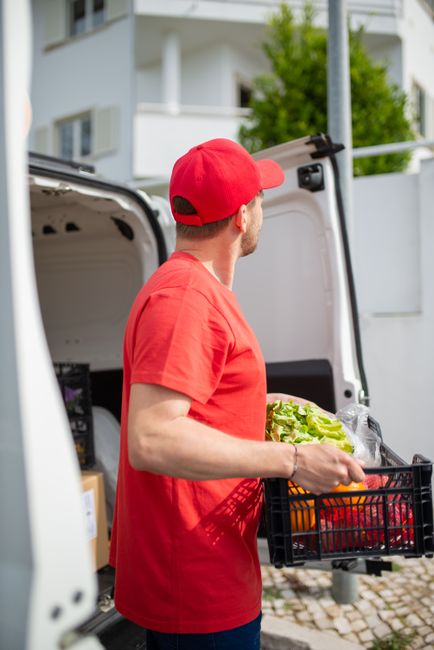 A man wearing a white t-shirt, jeans, and a black cap is sitting on the pavement next to an open delivery van during daylight, with the van loaded with multiple cardboard boxes of various sizes. The b