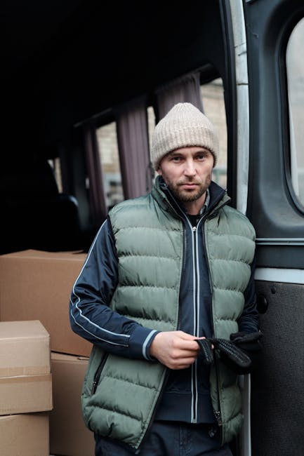 A man wearing a grey knitted beanie, a green padded front vest, and a navy blue jacket with light blue piping on the sleeves stands beside a dark grey van during a house removal process. The van's sliding door is open, revealing a partially visible back section filled with cardboard boxes of various sizes, likely containing household items for packing and moving. The man is holding a black strap or tie in his gloved hands, preparing to secure or unload items as part of the furniture transport and home relocation activities carried out by Man With a Van Kingsbury. The environment suggests an outdoor loading area or driveway under natural light, with the focus on the man's concentrated expression as he manages storage and transportation logistics for a relocation service.