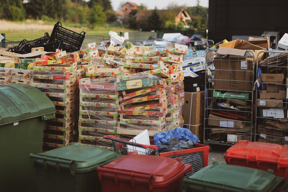 Outside a residential property, a collection of large wooden pallets stacked with empty cardboard boxes labeled 'fresh fruits' are arranged on a paved driveway or yard, indicating packing for a house removal. Adjacent to the pallets, there are several black plastic crates, some filled with cardboard and packing materials, along with metal shelving units loaded with boxes and miscellaneous items, possibly including small appliances or household goods. In the foreground, multiple plastic bins in green and red colors are visible, with some open and containing miscellaneous packing materials or items. A hand truck or trolley may be partially visible, suggesting the process of loading or unloading packing supplies. The environment is well-lit with natural daylight, and the background features a garden area with greenery and trees, indicating the prep work involved in a home relocation service, as carried out by Man With a Van Kingsbury, in relation to local housing or waste disposal rules.