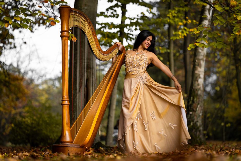 A woman dressed in an elegant golden gown with intricate embroidery and floral details is standing outdoors in a park during autumn, surrounded by trees with yellow and orange leaves. She is smiling a
