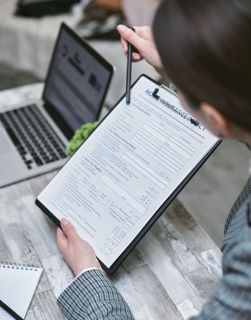 A person in a checked blazer is holding a clipboard with a detailed moving checklist or form, positioning it close to the camera, with a pen in their right hand pointing at the document. The setting appears to be indoors, possibly within a moving or packing environment, as a laptop displaying a document and a small, spiral-bound notepad are placed on a wooden table nearby. The background includes a blurred view of a workspace or home office, with natural lighting illuminating the scene. This image relates to packing, planning, or administrative tasks involved in house removals and local or office relocations, aligning with services provided by Man With a Van Kingsbury, who specialise in furniture transport and packing and moving logistics for home or office relocations.