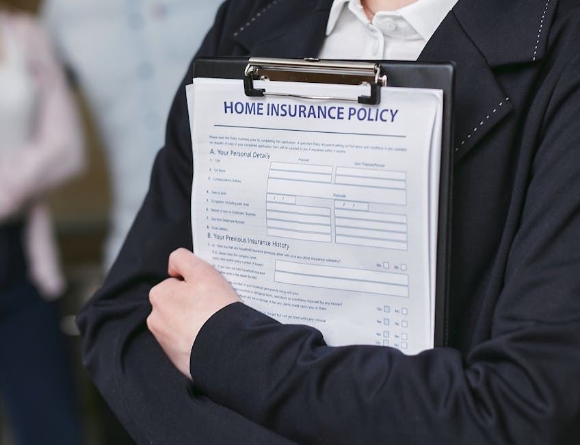 A person wearing a black blazer and white shirt is holding a clipboard with a 'Home Insurance Policy' document attached, which contains fields for personal details and insurance history. The individual is standing indoors in a well-lit environment, possibly during a house removal or moving consultation. The background is blurred but suggests an office or home setting, with another person partially visible on the left side. The scene illustrates the process of managing documents related to home relocation, with the clipboard held securely against the person's chest, indicating organization and preparation typical of professional removals services by Man With a Van Kingsbury.