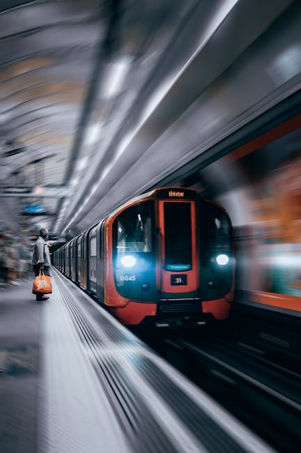Inside a modern underground station, a red and blue tube train is arriving, with its front illuminated and moving at speed. The station platform is visible on the left, where a person dressed in casual clothing is standing near the edge, carrying a small orange suitcase. The background shows blurred architectural elements and lighting, indicating motion and the bustling environment typical of London's public transport system. This scene reflects the logistics and transportation aspect of home relocation or moving services, highlighting the importance of reliable transit for furniture transport or packing and moving operations, as handled by companies like Man With a Van Kingsbury.