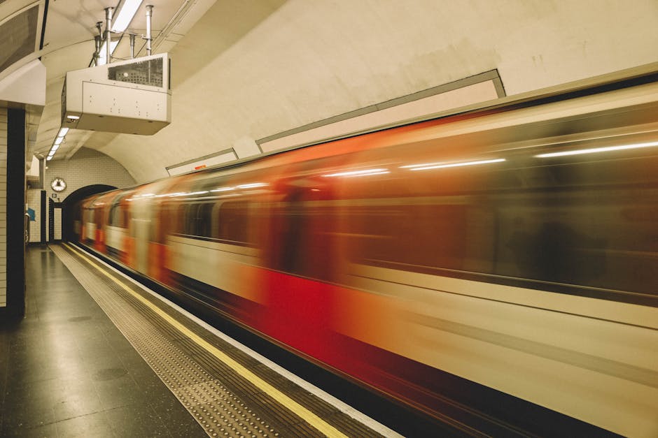 A moving train with a red and white exterior passing through the underground Kingsbury tube station in London, captured during a fast motion blur. The station platform in the foreground shows a black tiled floor with a tactile paving strip along the edge for safety. The station’s curved ceiling is painted in a light beige color, with visible lighting fixtures and ventilation pipes, and a large analog clock mounted on the wall near the tunnel entrance. Inside the station, a few cardboard boxes and wrapped furniture are temporarily stored close to the platform, with some items secured in protective plastic wrap and cardboard boxes. The movement of the train suggests an ongoing home relocation or furniture transport process managed by a professional moving service, such as Man With a Van Kingsbury, who may coordinate loading or unloading items from nearby vehicles or the station area.