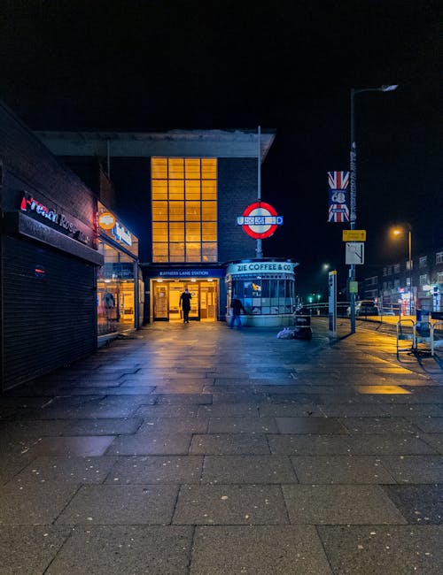 Inside a modern underground station, a red and blue tube train is arriving, with its front illuminated and moving at speed. The station platform is visible on the left, where a person dressed in casual clothing is standing near the edge, carrying a small orange suitcase. The background shows blurred architectural elements and lighting, indicating motion and the bustling environment typical of London's public transport system. This scene reflects the logistics and transportation aspect of home relocation or moving services, highlighting the importance of reliable transit for furniture transport or packing and moving operations, as handled by companies like Man With a Van Kingsbury.