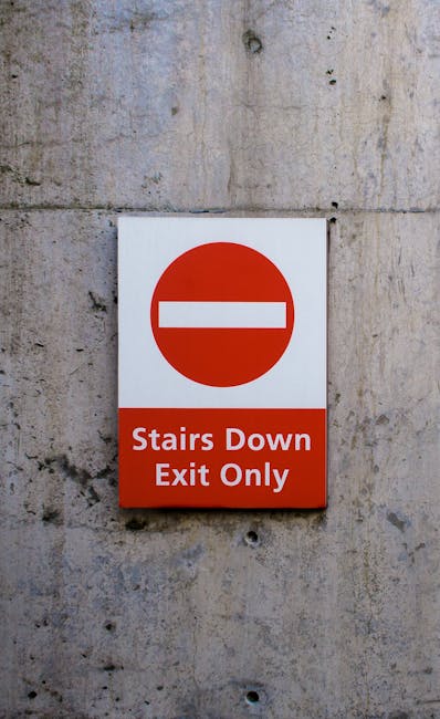 A rectangular road sign attached to a textured concrete wall, displaying a red circle with a white horizontal line in the center, indicating no entry. Below the circle, a red section contains white text that reads 'Stairs Down Exit Only'. The sign is positioned at eye level, suggesting it is intended for pedestrians or delivery personnel in a residential or commercial building. This context is relevant for house removals and packing and moving services, such as those offered by Man With a Van Kingsbury, particularly when navigating restricted access areas like staircases or confined stairwells during a home relocation. The lighting is natural or ambient, casting minimal shadows. The focus is on clear communication of access restrictions within a building’s interior environment, supporting safe and efficient furniture transport and loading procedures.