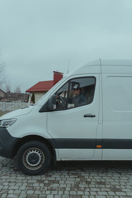 A white delivery van parked on a cobblestone driveway with a man sitting in the driver's seat, dressed in a grey jacket and cap. The van is positioned in front of a red-roofed house with white fencing and a garden visible in the background. The side door of the van is closed, and the vehicle appears ready for home relocation or furniture transport services. The scene is outdoors during daytime under a cloudy sky, with the van representing a professional moving or removal service, as advertised by Man With a Van Kingsbury, in the context of house removals and packing and moving processes.