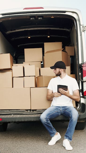 A man with a short beard and wearing a white t-shirt, light blue jeans, white sneakers, and a black cap is sitting on the pavement beside the open rear of a white cargo van. Inside the van, numerous uniformly sized cardboard boxes are stacked, some placed directly on the van floor while others are arranged on top. The boxes are sealed with packing tape and appear to be part of a home relocation or furniture transport process managed by Man With a Van Kingsbury, indicated by the professional packing and loading setup. The van's interior shows a dark, spacious cargo area suitable for moving household items, with some clothing or padding materials possibly used for protection during transport. The scene is outdoors in natural daylight, with the focus on the loading or unloading phase of the furniture transport service. This image illustrates packing, furniture transport, and moving logistics involved in house removals, aligned with services explained on the NW9 removals and man with a van services page at manwithavankingsbury.co.uk.