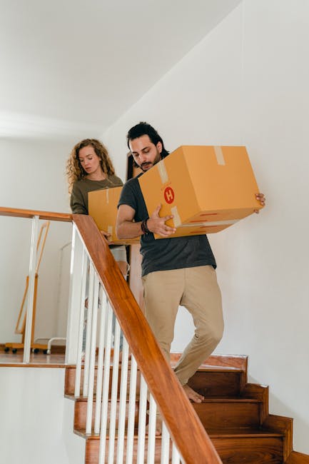 A man and a woman are carrying cardboard boxes, with the man holding a large box featuring a red 'H' logo, during an indoor home relocation process. They are descending a wooden staircase with white spindles, using caution with the boxes. The setting appears to be inside a residential property with neutral-white walls and natural lighting. The man is dressed in a dark t-shirt and beige trousers, while the woman wears a brown top. This scene depicts the packing and furniture transport phase typical of house removals, with the manwithavankingsbury.co.uk offering professional removals services for efficient moving logistics and packing. The image emphasizes the careful handling of boxes and the use of protective materials like cardboard during the loading process within the home environment.