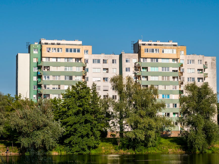 A multi-storey residential block in Queensbury with a grey facade accented by green balconies, situated behind a line of mature trees and greenery near a body of water, under a clear blue sky. The building appears to be part of a typical home relocation setting, with windows and balconies indicating occupants' living spaces. This scene illustrates the environment where professional removals services from Man With a Van Kingsbury might be undertaken, highlighting the importance of organized packing, furniture transport, and loading processes during a house move or apartment relocation within the Queensbury estate.