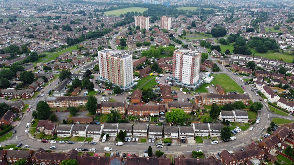 Aerial view of a residential area showing a mixture of housing types, including two prominent high-rise apartment buildings with white and red exteriors situated amidst a neighborhood of smaller terraced and detached houses. The scene features tree-lined streets, parked cars along the curbs, and patches of green lawns and communal gardens. The surrounding landscape includes open fields and additional housing further in the distance. Visible in the foreground is a street with cars parked parallel, leading into the estate, which appears to be part of a home relocation or moving logistics operation. The image captures the organized layout of the flats and the adjacent houses, emphasizing the urban residential environment typical for house removals and furniture transport services, as offered by Man With a Van Kingsbury.