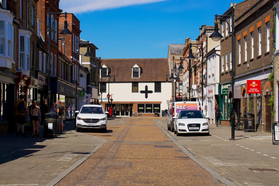 A view of a busy street in Kingsbury with shops and pedestrians. In the center, a white van is parked on the cobblestone street, with a man with a black jacket and cap loading or unloading items nearby. To the right, a white Audi car is parked, and a delivery van with a pink logo is adjacent. On the left, a group of people stands outside shops with large glass windows and mannequins, while black street lamps line the sidewalks. The buildings are a mix of brick and painted facades, and the sky is clear and blue, indicating a bright day, suitable for home relocation or furniture transport activities. This scene reflects the logistical aspect of house removals, with vehicles and pedestrians involved in moving or packing processes, supported by services from Man With a Van Kingsbury.