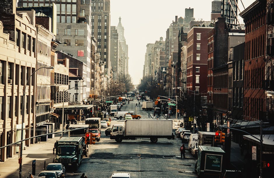 A broad city street scene in daylight showing multiple multi-storey buildings with varied architectural styles on either side, housing retail shops and offices. The street is busy with parked and moving vehicles, including delivery trucks, vans, and cars, as well as pedestrians walking on the pavements. In the foreground, a white medium-sized van is positioned on the street, possibly involved in a home relocation or furniture transport. The vans and trucks are complemented by traffic lights and street signage overhead. The lighting suggests a late morning or early afternoon setting, with sunlight casting shadows across the street. The scene depicts urban logistics, likely involving the loading or unloading of household belongings, fitting within the context of house removals and moving services provided by Man With a Van Kingsbury, emphasizing urban moving operations and moving logistics within a busy city environment.